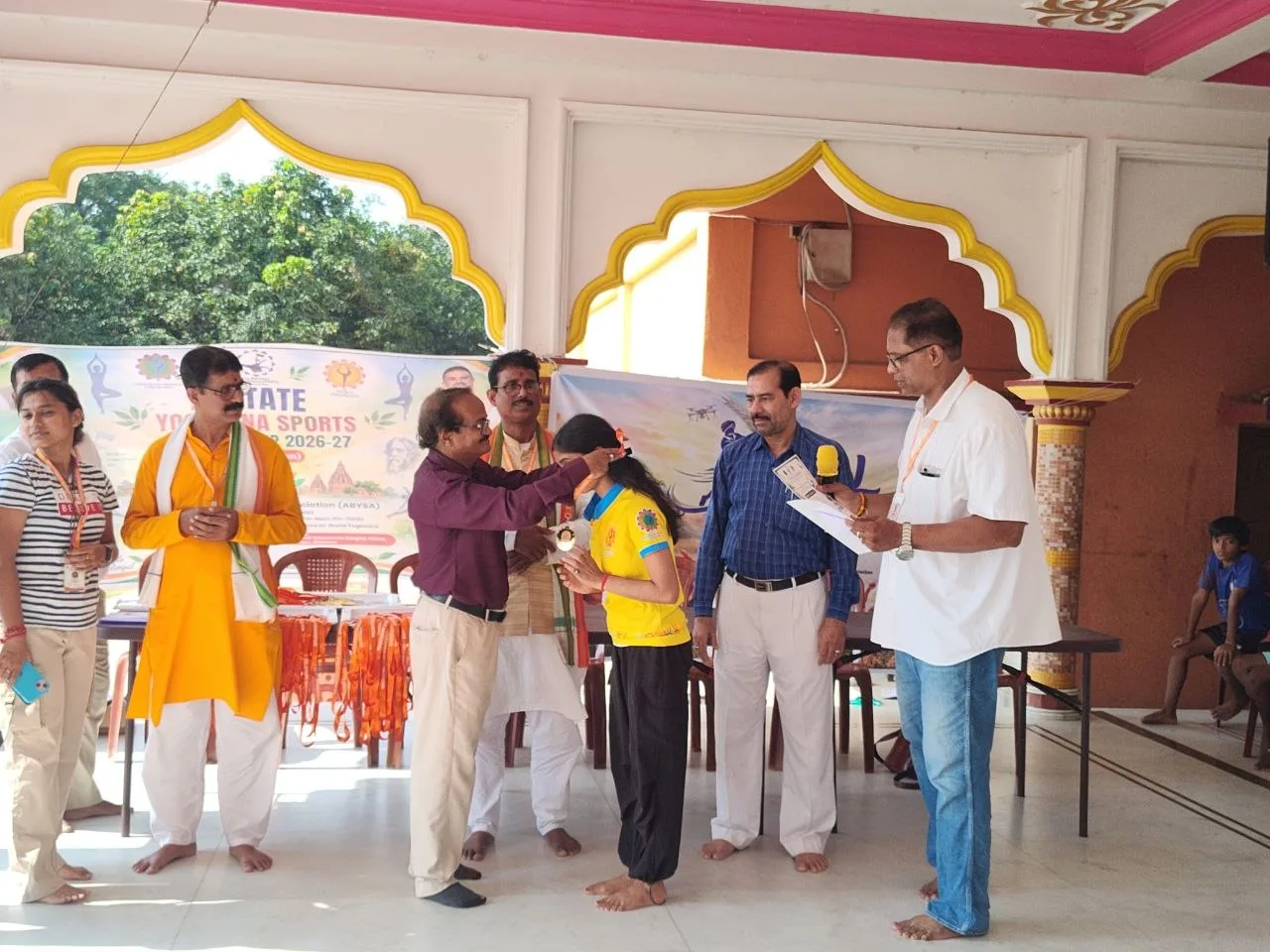 Officials stand with an athlete in yellow as a certificate is held in the hall.