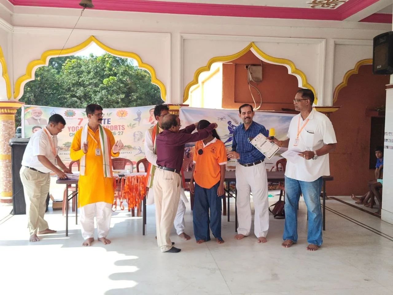 Officials place a medal over an athlete in an orange shirt while a certificate is held beside him.