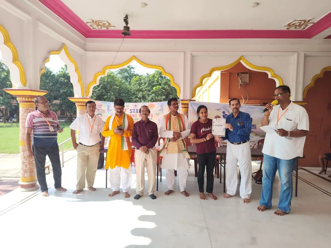Officials stand with an athlete in a yellow saree as a certificate is held in the hall.