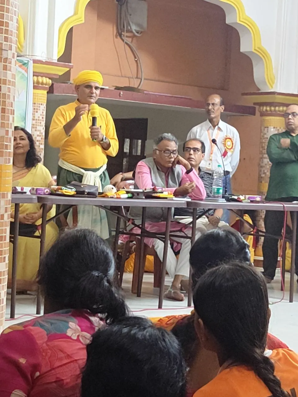 Sourabh J. Sarkar, in yellow, stands with a microphone beside the long table.